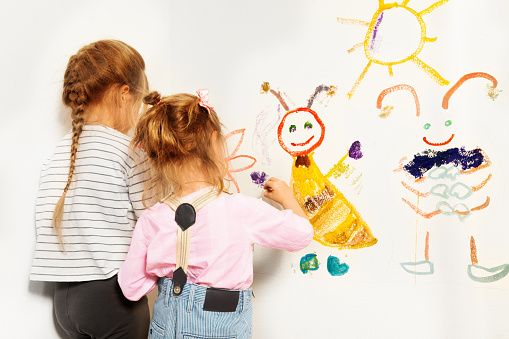 Two little painters, preschool girls drawing funny picture at the wall, isolated on white