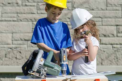 Caucasian boy drilling while his sister is holding a hammer, on the workbench. The boy wears a yellow hard hat and the girl a white hard hat. They are outside in the backyard during a day of summer.