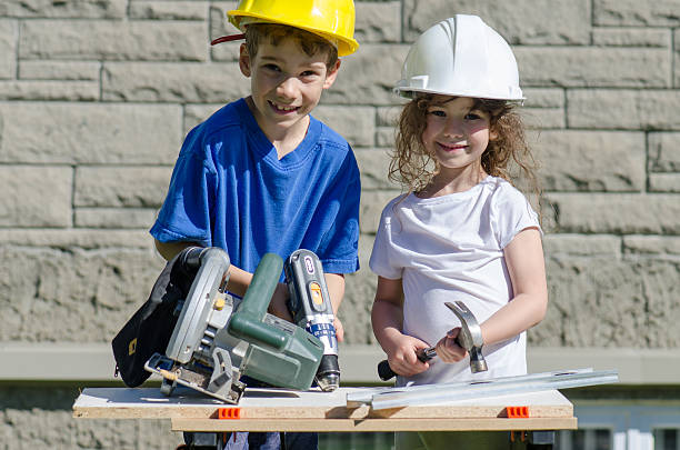 Little boy and girl playing with big tools outside Caucasian boy drilling while his sister is holding a hammer, on the workbench. The boy wears a yellow hard hat and the girl a white hard hat. They are outside in the backyard during a day of summer. girl with a hammer stock pictures, royalty-free photos & images