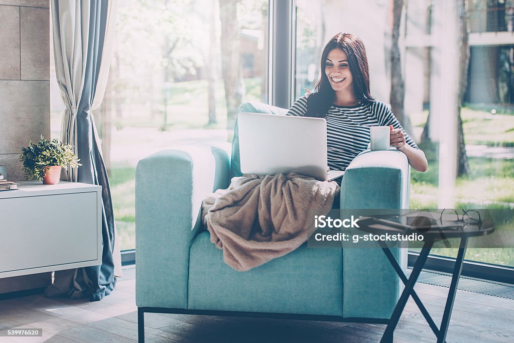 Enjoying time at home. Beautiful young smiling woman working on laptop and drinking coffee while sitting in a big comfortable chair at home Domestic Life Stock Photo