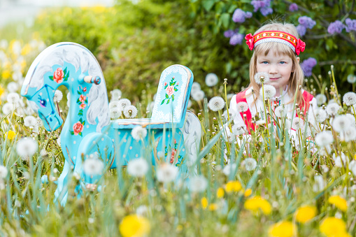 Happy blonde little girl dressed in Slavic traditional costume. The cute little girl is sitting  on a green meadow with dandelions. She is looking at the camera. The wooden rocking horse painted with patterns on foreground. Shooting at sunny summer day