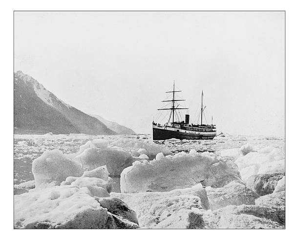 Antique photograph of of a vessel sailing in the Glacier Bay Basin (Alaska, USA) during the 19th century. In the picture the surrounding mountains and glaciers and the ship breaking the ice in the freezing waters of the Ocean, surrounded by ice plates and small iceberg 