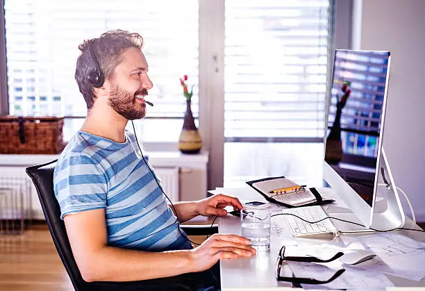 Man sitting at the desk working from home on computer, talking on the phone, headset on head, smiling