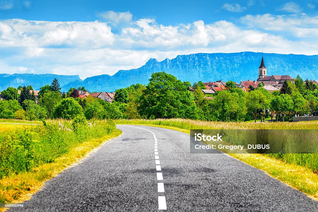 Empty countryside road with old french village and Alps mountains Horizontal composition HDR vibrant color photography of a typical small old French village of Contrevoz, in Bugey mountains in Ain, Auvergne-Rhone-Alpes region in France (Europe) slightly elevated, with its apparent steeple. This picture was taken next to an asphalt countryside empty road. We distinguish the mountains of the Alps in background with a blue sky with some clouds. France Stock Photo