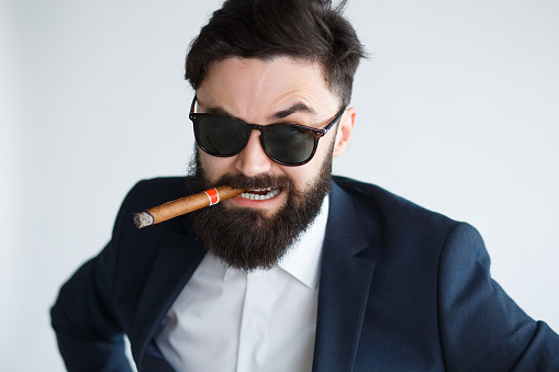 Closeup portrait of boss man in black suit and sunglasses smoking cigar on white background. Angry bearded businessman