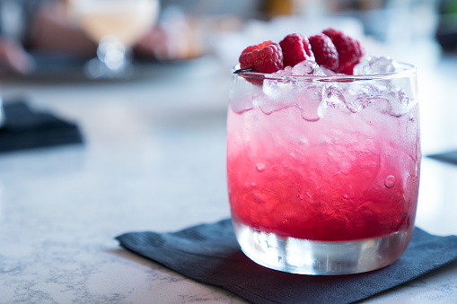 A photograph of a tumbler glass filled with crushed ice and a Vodka and Raspberry mix, with a skewer of 4 raspberries across the top ot the glass. The tumbler glass with the Vodka Raspberry mix is sitting on a napkin on a marble bar. The glass with the Vodka Raspberry mix is in focus with marble bar top and everything in the background falling out of focus.