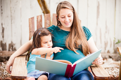 Attractive young pregnant woman reads to her daughter in the back yard.
