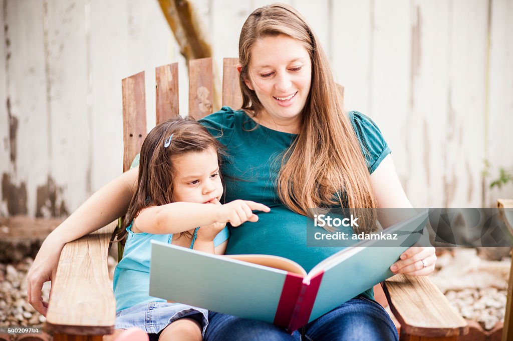 Pregnant Woman Reading to Daugther in Backyard Attractive young pregnant woman reads to her daughter in the back yard. Rural Scene Stock Photo