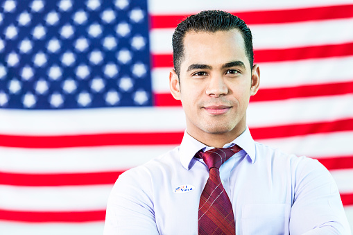 Handsome Hispanic man standing in front of an American flag. He is wearing a button up shirt and red tie. He is also wearing an :i voted\