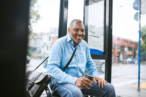 A business man waits at a city bus stop with a coffee in hand and a nice leather messenger bag over his shoulder.  He smiles as he waits.  African American ethnicity; 50-60 years old.  Shot in Portland, Oregon.  Horizontal image.