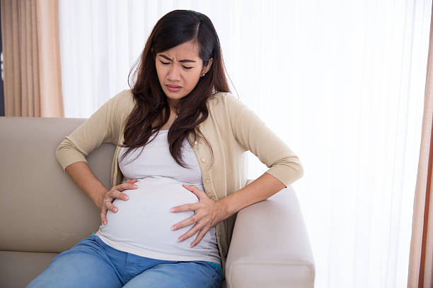 азиатские с беременная женщина. �сидя на ее диване, боль в животе - muscular contraction стоковые фото и изображения