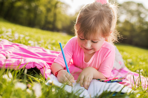 Little girl relaxing on a blanket outdoors and drawing in a notebook.