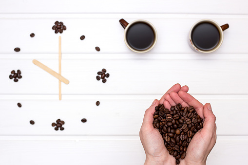 Coffee beans in the hands above th table with clock and two cups of coffee
