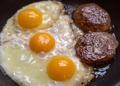 A close view of eggs and sausage frying in a skillet next to a window for natural light.