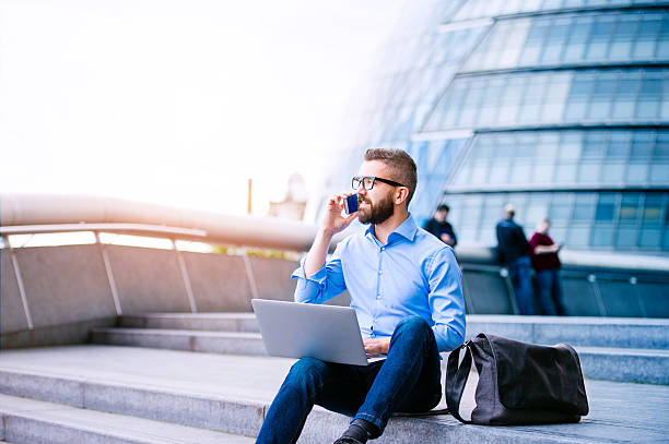 Manager with laptop and smart phone, London City Hall Handsome hipster manager sitting on stairs on sunny day, working on laptop, talking on a smart phone, London, City Hall blue business stock pictures, royalty-free photos & images