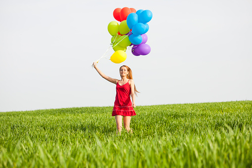 Cheerful young woman holding a bunch of colorful balloons and running in the grassland, hand raised. Wears red dress.