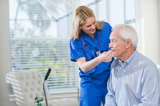 Home healthcare nurse helping elderly man with oxygen Home healthcare worker helping a senior man with his portable oxygen tank. She is standing beside him in blue scrubs, adjusting the tubing. oxygen photos stock pictures, royalty-free photos & images