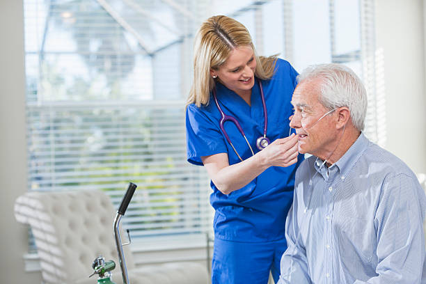 Home healthcare nurse helping elderly man with oxygen Home healthcare worker helping a senior man with his portable oxygen tank. She is standing beside him in blue scrubs, adjusting the tubing. medical oxygen stock pictures, royalty-free photos & images