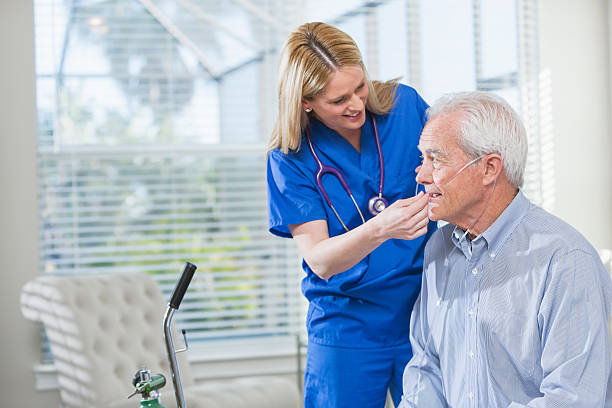 Home healthcare nurse helping elderly man with oxygen Home healthcare worker helping a senior man with his portable oxygen tank. She is standing beside him in blue scrubs, adjusting the tubing. o2 stock pictures, royalty-free photos & images