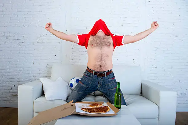 young man watching football game on television celebrating goal crazy with jersey on his head jumping on sofa couch at home with ball holding  beer bottle eating pizza excited