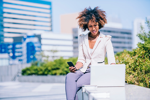 Young black woman is using her laptop. She is focused, holding her mobile phone and looking at her laptop. She is sitting on a bench with laptop in front of her in a park. Green trees and business buildings in the back.