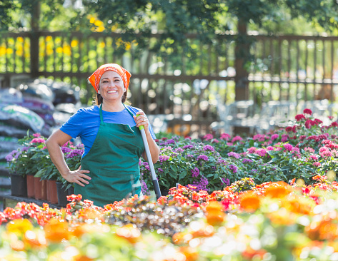 An Hispanic woman in her 50s working in a plant nursery, standing in the middle of tables of potted flowers with her hand on her hip, holding a broom.  She is smiling and looking confidently at the camera.