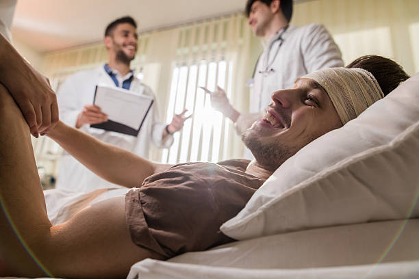 Happy patient lying in hospital bed and holding hands with unrecognizable person while two doctors are communicating in the background.