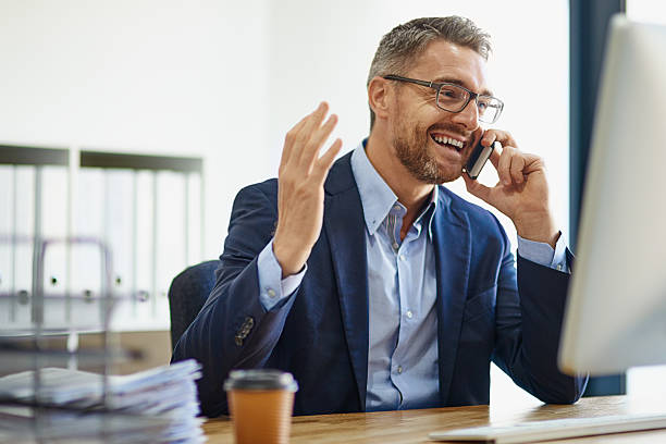 Expanding his empire using technological resources Shot of a mature businessman using his phone and computer at his desk good news stock pictures, royalty-free photos & images