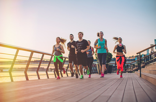 Group of young people competing in a race. Young men and women running on riverside promenade at sunset. They are wearing sport clothing.