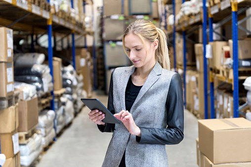 Portrait of a pensive young woman supervisor working on-line in warehouse. Young blond woman standing at distribution warehouse and wearing elegant suit. Industrial boss examining the stocks. Large distribution storage in background with racks full of packages, boxes, pallets, crates ready to be delivered. Logistics, freight, shipping, receiving.