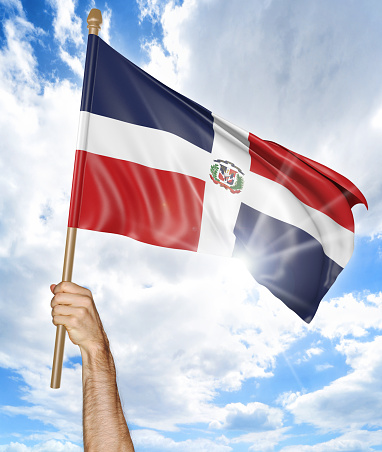 Person holding the national flag of the Dominican Republic high in the air against a bright sky and sun rays.