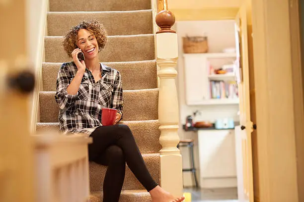 a woman sits on the stairs of her home and  and is laughing on the phone  . She is smiling happily on her landline holding a cup of tea . In the background, a hallway and kitchen are visible.