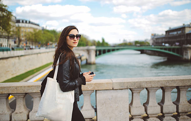 Vintage toned image of a young Parisian woman walking the Paris city center, by the river Seine on a beautiful spring or early summertime day. She is wearing a black leather jacket, carrying a tote bag, on her way to work. Paris, lifestyle, tech, street style fashion concepts.