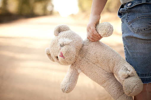 Color stock photo of a little runaway girl holding an old teddy bear at the side of a dirt road in the rural country.