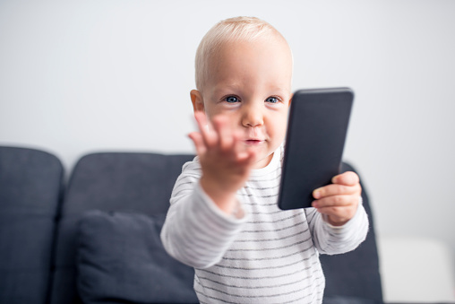 Cute young little boy holding smart phone. He is sitting a sofa and is focused on mobile phone.