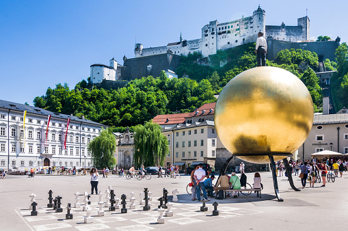  Salzburg, Austria - May 26, 2016: Visitors relax on benches in the shade of a modern statue at Kapitelplatz at the historical center of Salzburg. The  sculpture of a golden sphere, called Sphaera, with a human figure standing on top was created by sculptor Stephan Balkenhol in 2007. A large chess board is nearby, and Salzburg Castle rises above.
