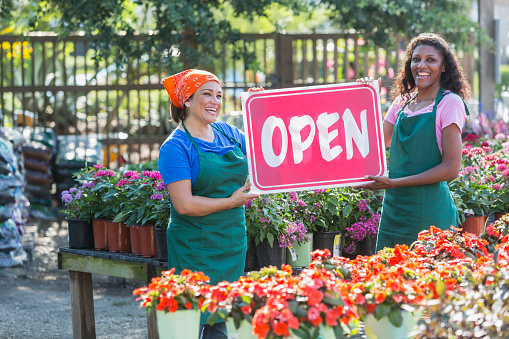 Two multi-ethnic mature women standing in the middle of a plant nursery, surrounded by potted flowers, holding an large, red, OPEN sign. This is the grand opening of their new small business.