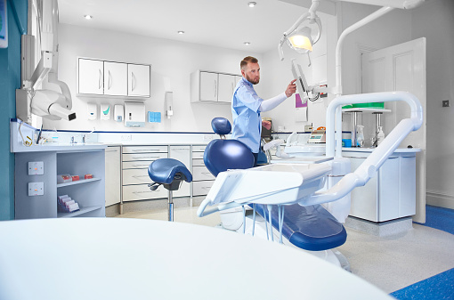 a mid adult male dental nurse or dentist in a  modern brightly lit dental surgery . The room is bright , modern and minimal in it's layout. He appears as he dials up an x-ray on his screen .