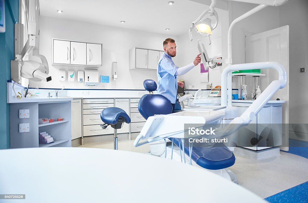 young male dental nurse or dentist a mid adult male dental nurse or dentist in a  modern brightly lit dental surgery . The room is bright , modern and minimal in it's layout. He appears as he dials up an x-ray on his screen . Dentist Stock Photo