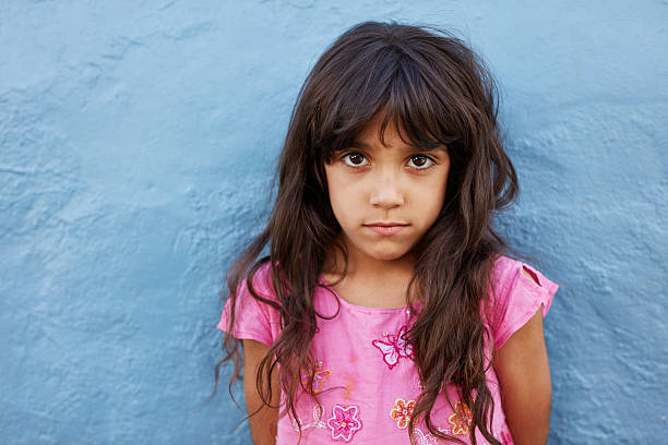 Innocent little girl standing against blue wall Close up portrait of innocent little girl standing against blue wall, she is looking at camera with serious expression on her face. blank expression stock pictures, royalty-free photos & images