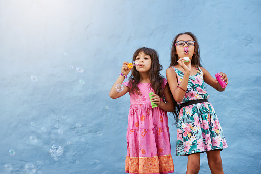 Portrait of adorable little girls blowing soap bubbles against blue wall. Two young girls playing together.