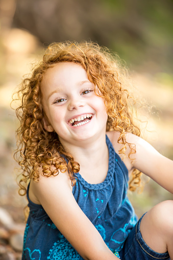 Close-up of a young girl with curly red hair sitting on the bank of a river laughing and looking at the camera. Taken on a summer day in Minnesota along the Mississippi River.