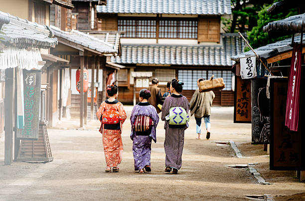 Rural scene in old Japanse village with wooden houses Two young and one elderly Japanese women wearing kimonos and yukatas while walking in traditional Japanese village, followed by Japanese peasants. Image taken with Nikon D800 and developed from RAW in XXXL size, in TOEI studios in Kyoto, Japan. yukata photos stock pictures, royalty-free photos & images