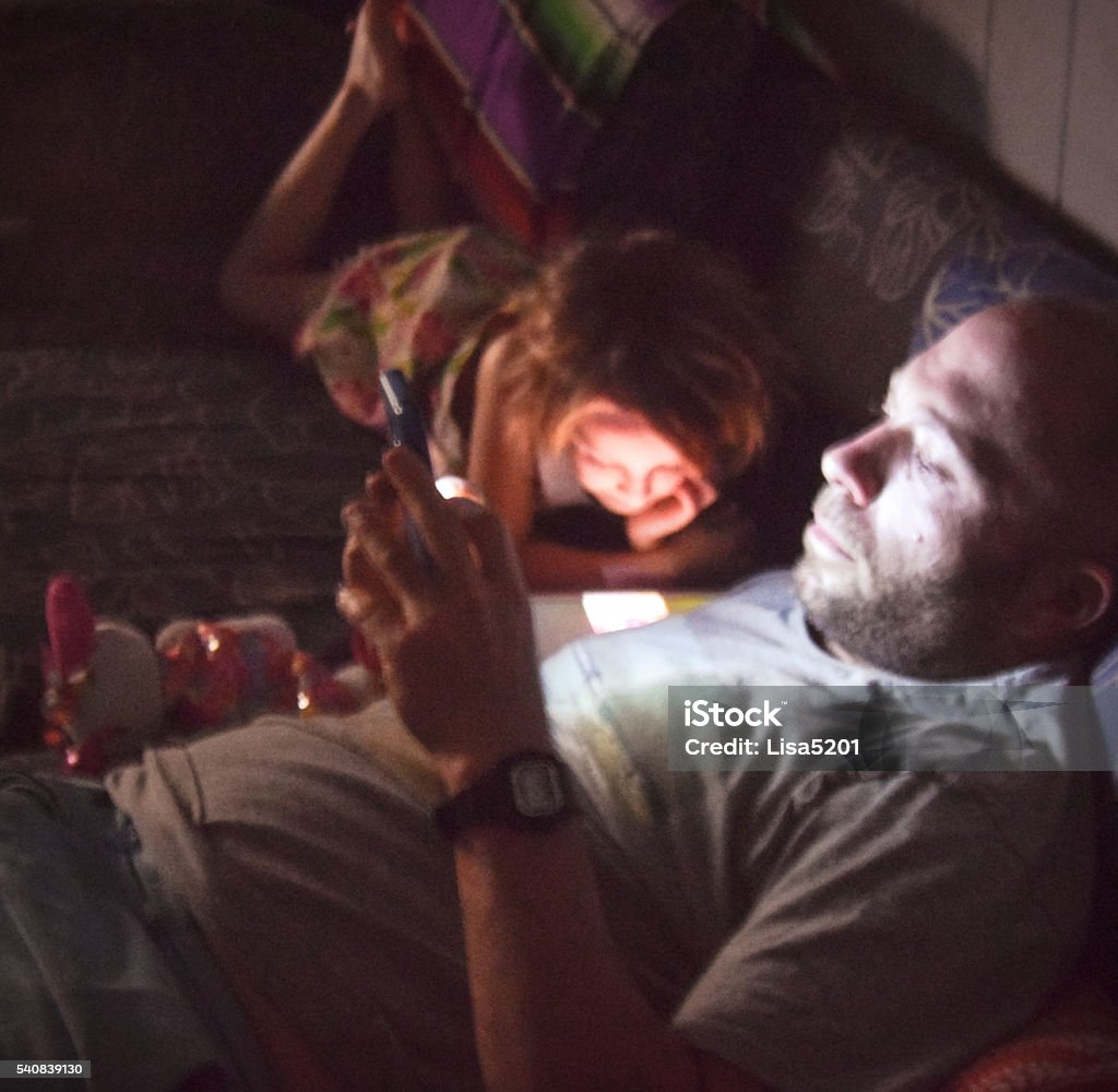 Screen time Father on his phone illuminates his face, daughter in the background on a device as well Screen Time - Concept Stock Photo