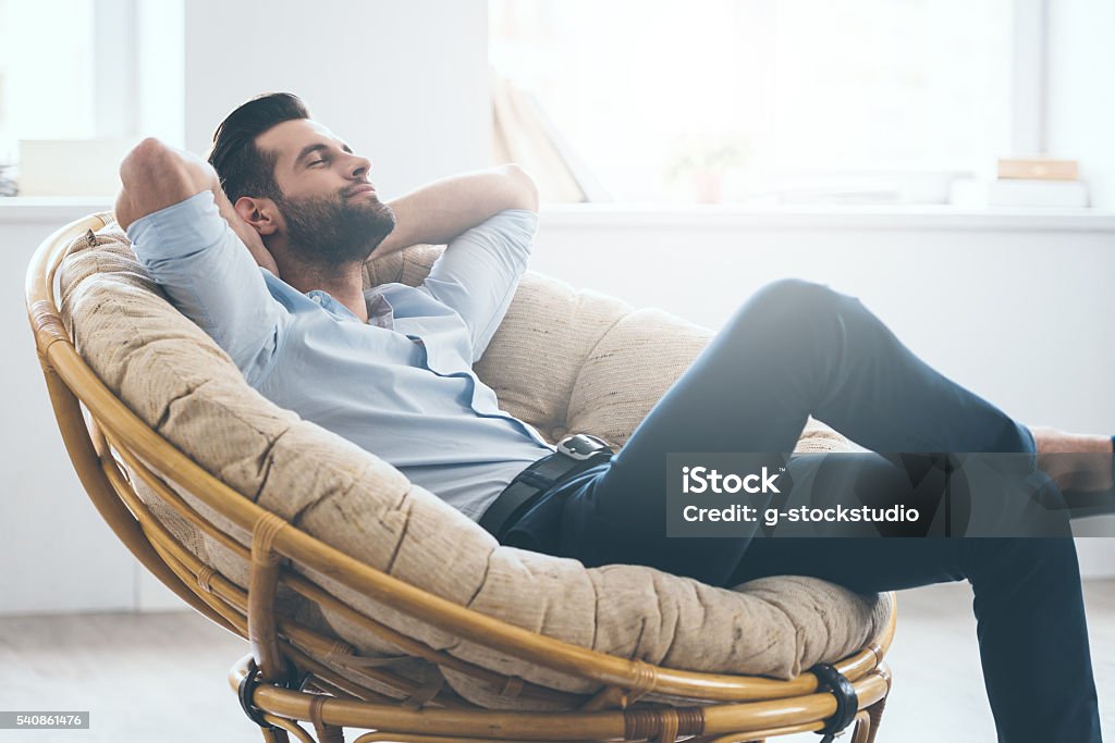 Total relaxation. Handsome young man keeping eyes closed and holding hands behind head while sitting in big comfortable chair at home Relaxation Stock Photo