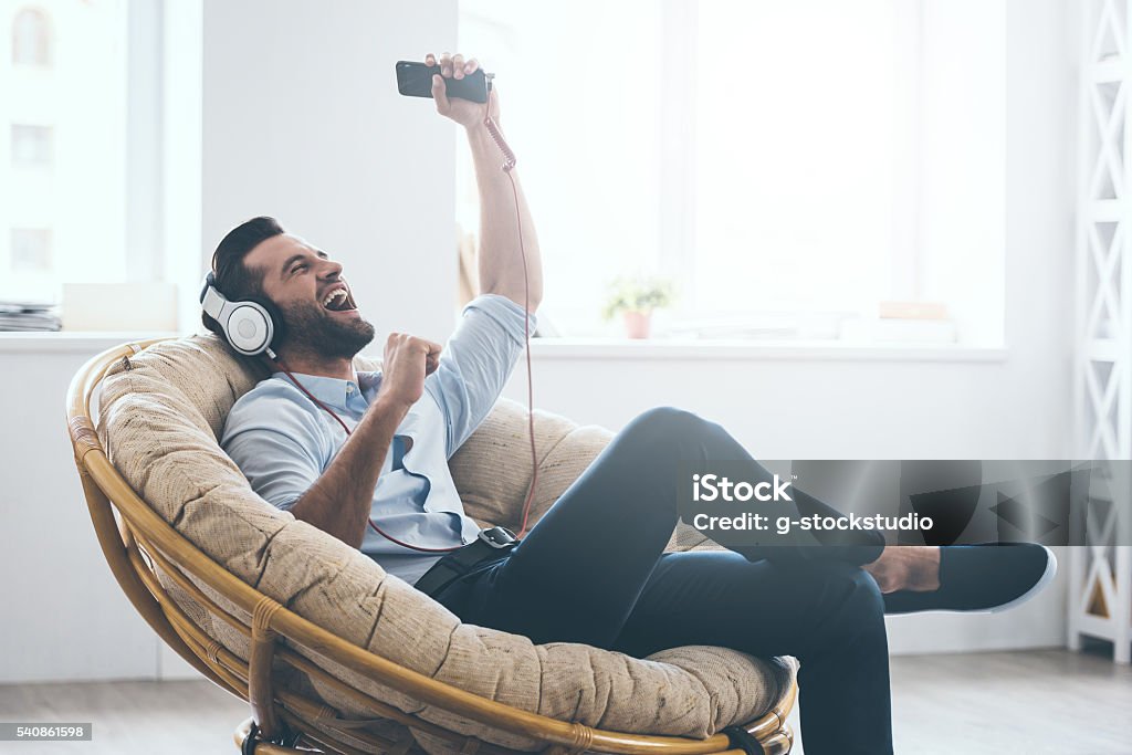Time to relax. Handsome young man in headphones gesturing and keeping eyes closed while sitting in big comfortable chair at home Men Stock Photo