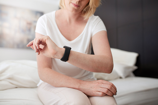 Cropped shot of a woman sitting on bed checking time on her wrist watch, she is in bedroom at home.