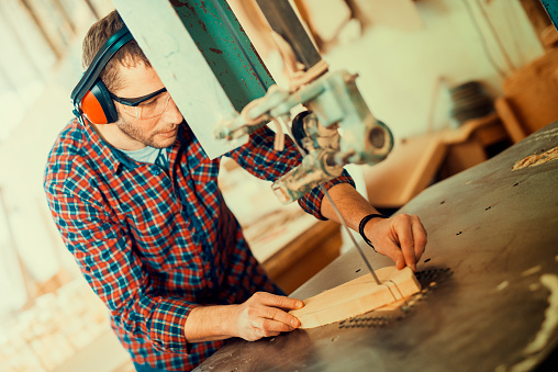 Close up of a young carpenter at work.He is using a bandsaw, also known as a jigsaw or a scroll saw, cuts through a piece of wood.