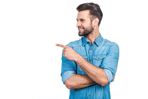 Happy young handsome man in jeans shirt pointing away and smiling while standing against white background