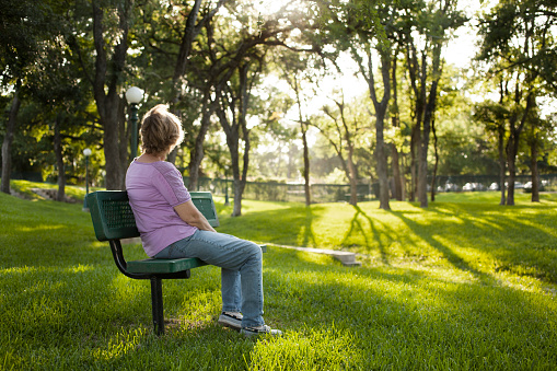 Rear view of one mature woman sitting on a park bench in the summer season.  Bright sunny day with lush green grass and trees.  The woman gazes off into the distance as she relaxes on a beautiful day.  Solitude, lonliness, contemplation.  She has short blond hair and wears a purple shirt and jeans.   Copyspace to right in this tranquil nature scene.